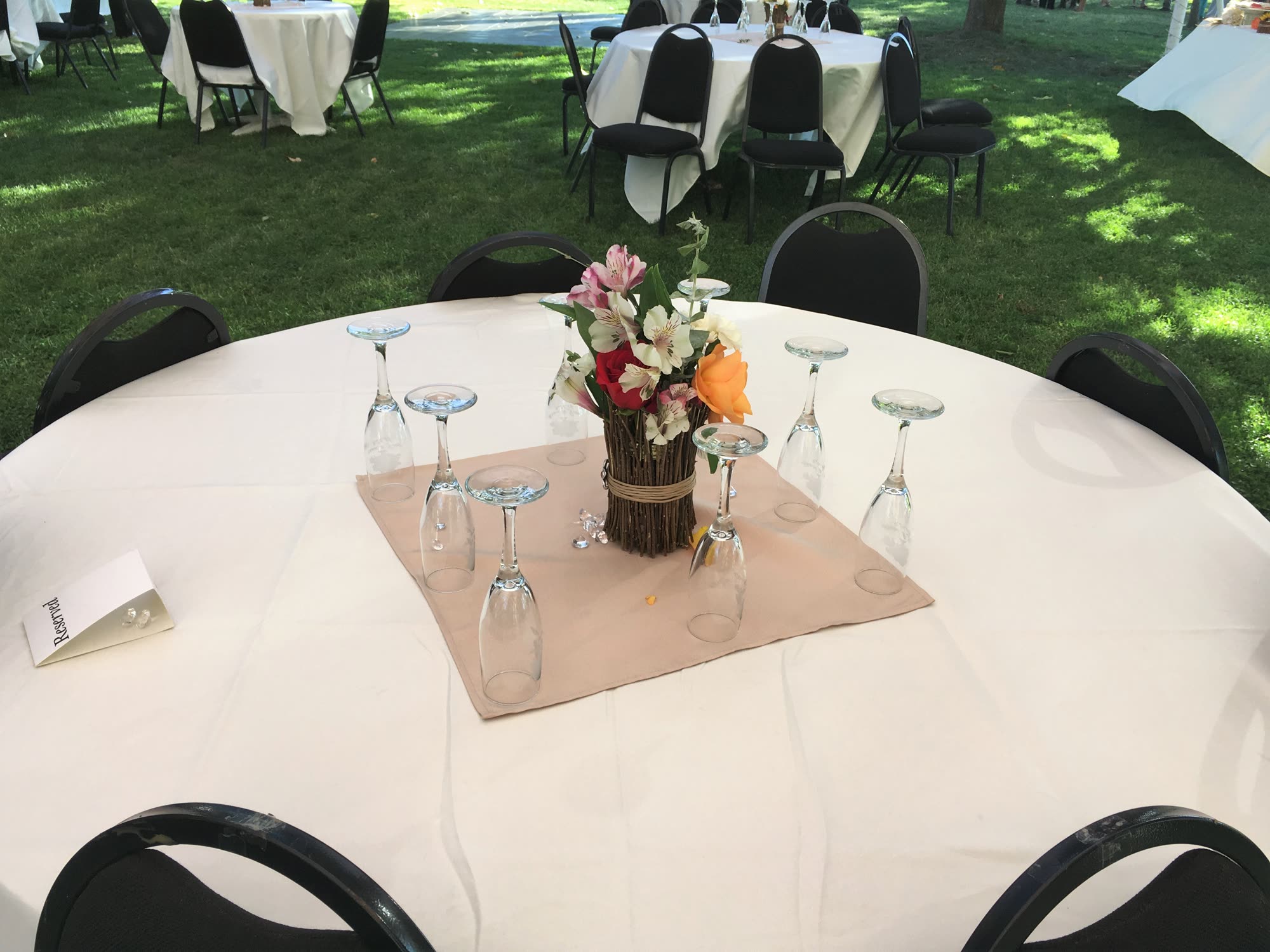 A round guest table in dappled shade, glasses upturned around a twig-wrapped floral centrepiece