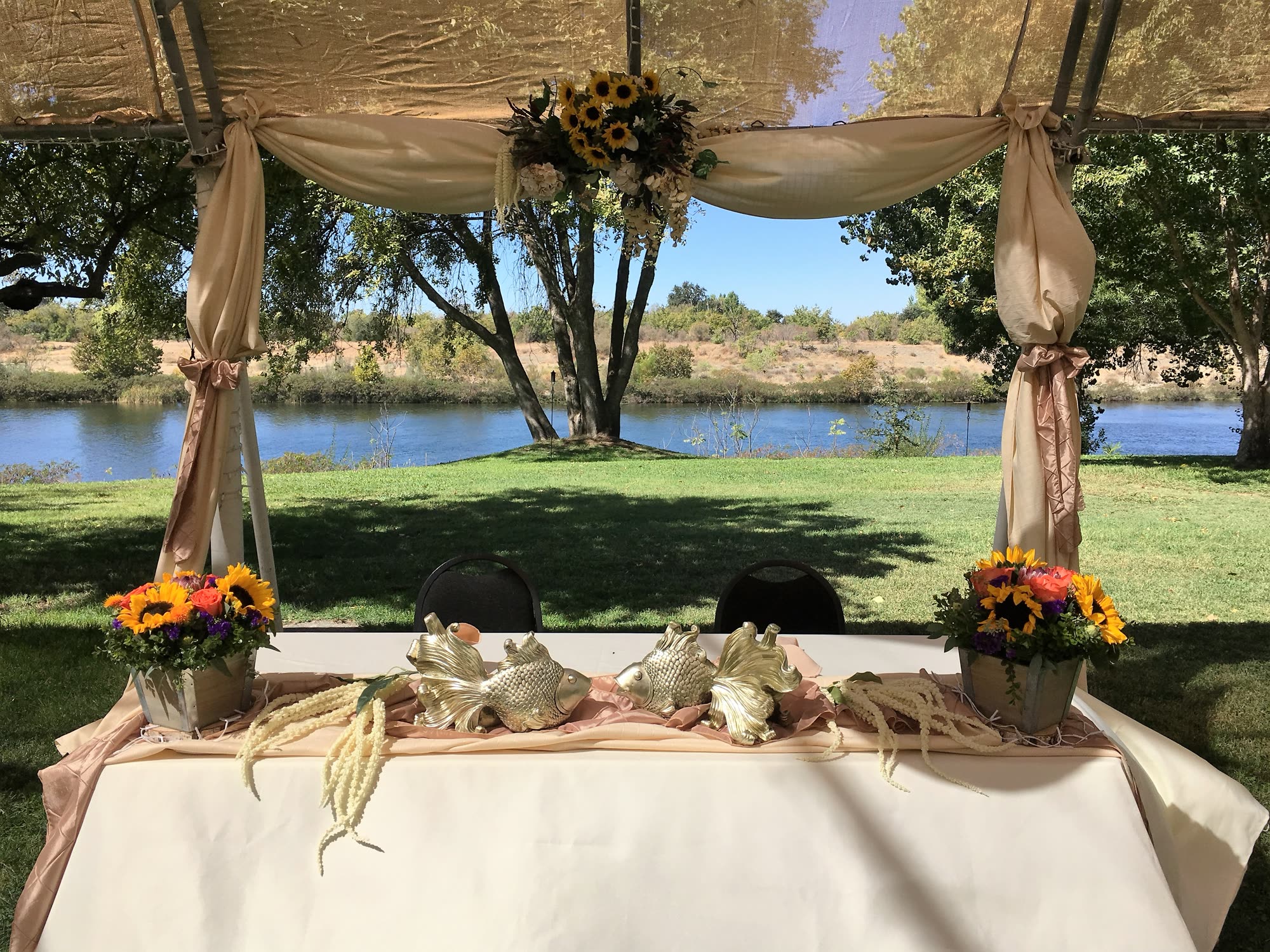 Head table dressed with sunflowers and gold fish sculptures, framed by drapery and the river